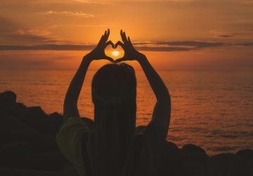 Lady holding hands in heart over sun at sunset